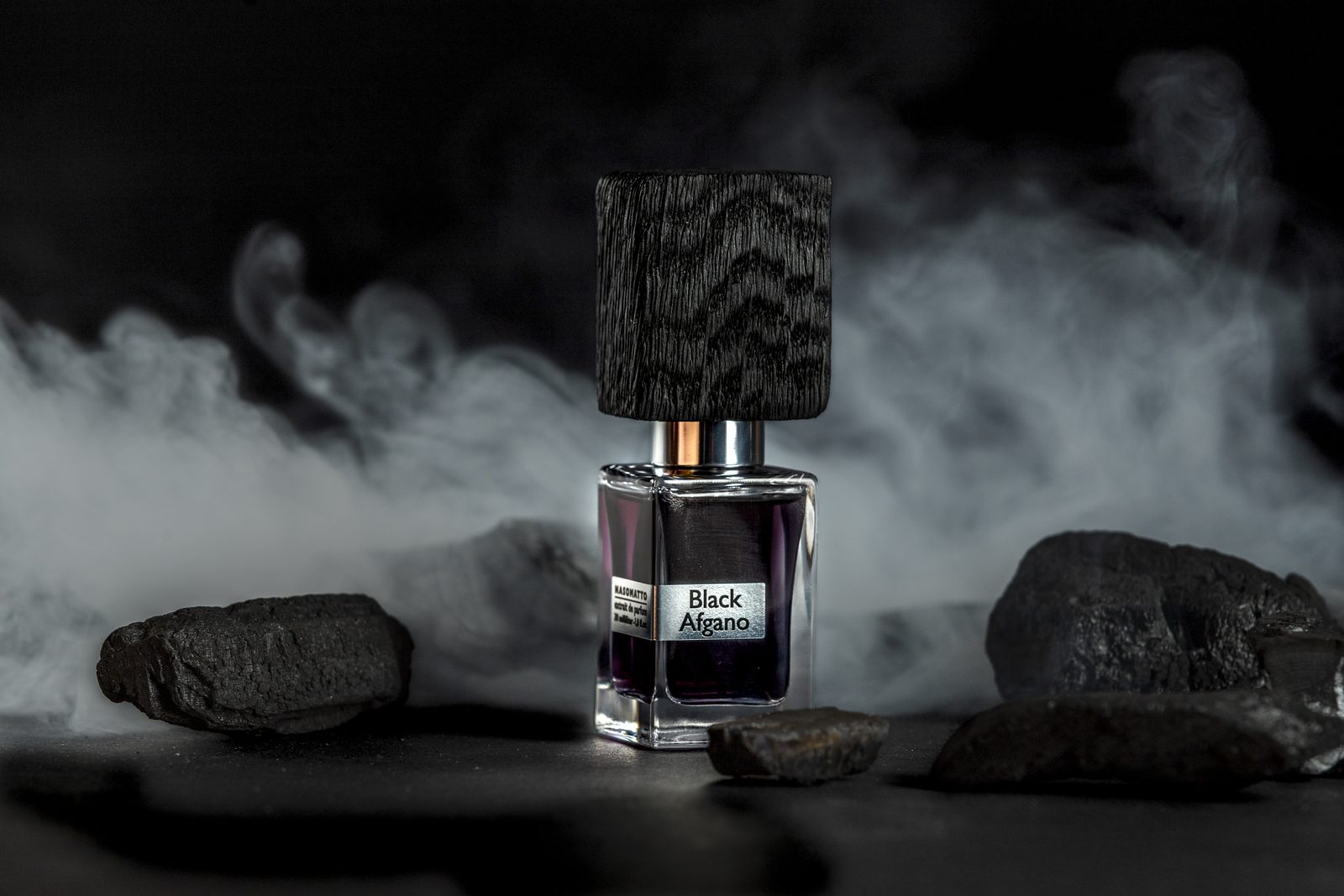 Still life di una boccetta di profumo tra carboni e fumo, luce controllata in studio